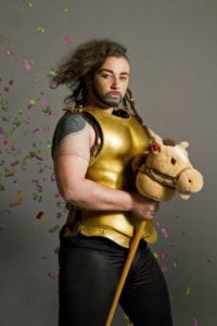 An androgynous person dressed in plastic gold armour poses with a hobby horse while a glitter canon goes off behind them