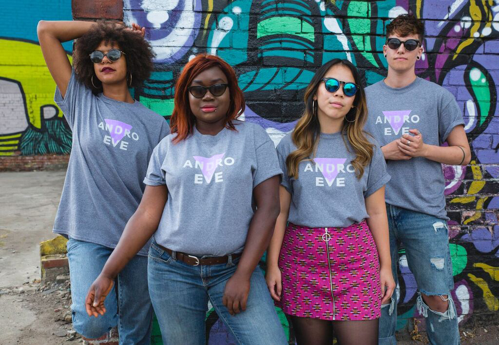 4 people stand with sunglasses on wearing the grey marl Andro and Eve Logo Tee. Two of the people are Black women wearing blue jeans, one person is an Asian woman wearing a pink skirt, and another person is a transmasculine person wearing blue ripped jeans. 