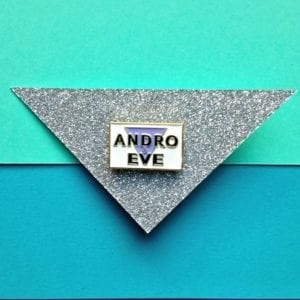 A rectangle white enamel pin badge with gold edging. The Andro and Eve logo, a lilac inverted triangle has the words Andro and Eve on top with an ampersand symbol for and. The badge is mounted on a glittery silver piece of card placed upon a turquoise surface.