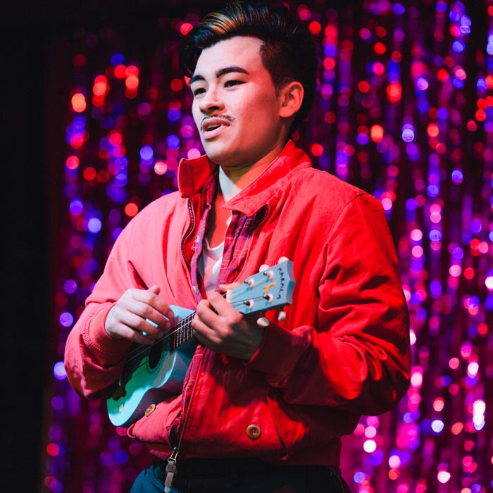 an East Asian drag king, Sigi Moonlight strums a small banjo onstage. He wears a red shirt and black trousers and has short black hair with a bleach blonde streak at the front