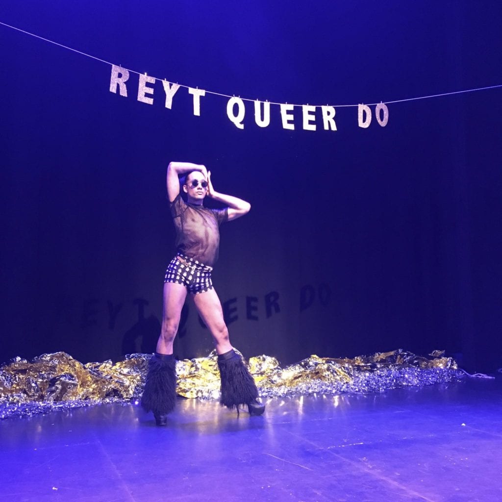 A tall, Black dancer with their hair tied back stands posing on a stage with their arms framing their face. They wear black and white hot pants and a mesh black top and black furry boots. Behind them is gold letter bunting spelling out Reyt Queer Do