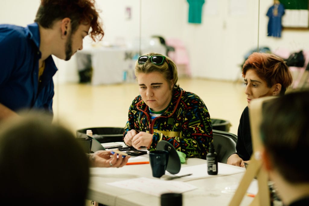 Lucy leans over a table to help share drag king makeup skills to two participants who have painted on some thick eyebrows