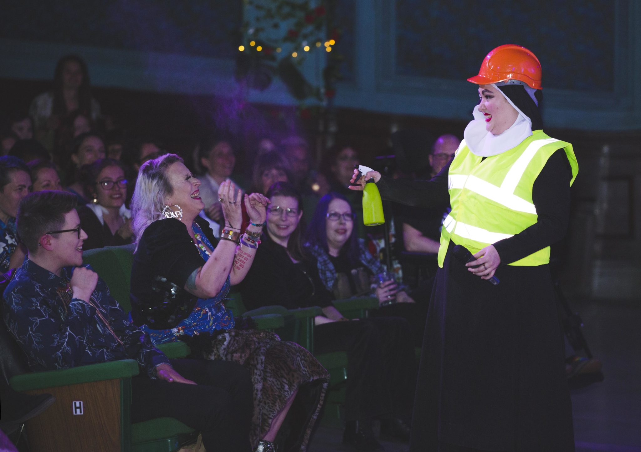 Sister Mary Berry, of drag act, Shesus and the Sisters, wears a hard hat and high vis vest and is squirting an audience member with mist from a bottle looking cheeky.