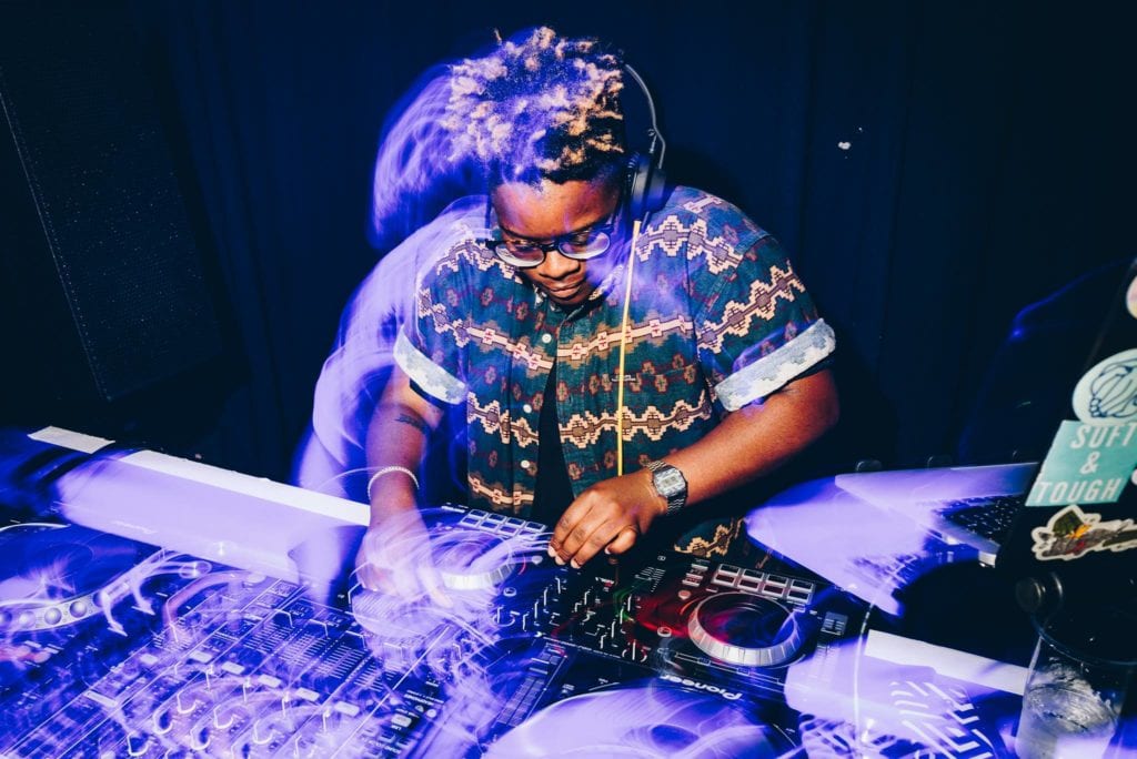 A Black non binary person DJ's on some decks. They are lit by purplelighting and wearing a green, geometric patterned shirt and glasses.