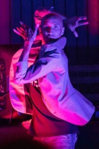 A Black dancer strikes a classic old school Vogue pose on a runway. His hands are around his face and he wears a light suit jacket, jeans and T Shirt. He is lit dramatically by pink and purple lighting. 