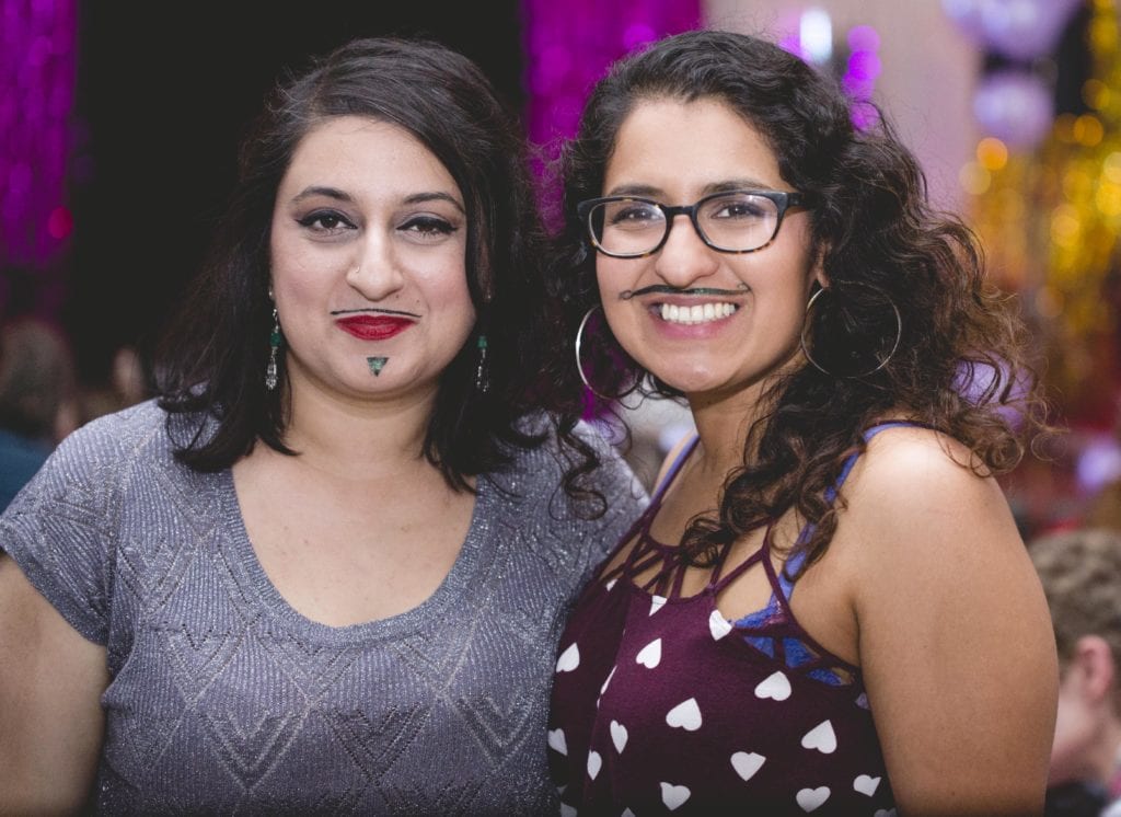 two women, one south Asian with shoulder length brown hair and the other middle eastern with long curly hair stand smiling with their arms round one another. They both have moustaches drawn on their face with eyeliner.