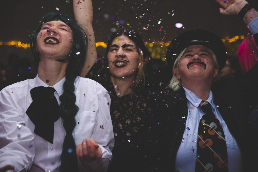 three white people look upwards with smiles and eyes closed as they enjoy glitter being thrown over theirselves in a dark party setting