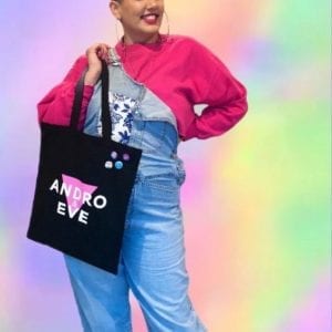 a plus sized Indian woman with a shaved head models the black andro and eve cotton tote with pink and white logo design. She wears a hot pink jumper under a pair of blue denim dungarees with pale pink trainers and is surrounding by a rainbow cloud.