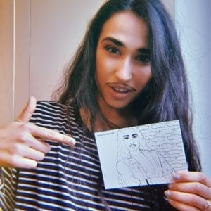 Christian Adore holds a postcard with the image of himself hand drawn in black and white on it. Christian is an olive skinned drag king with long black hair, black eyeliner and thin black moustache. He wears a black and white striped t shirt.