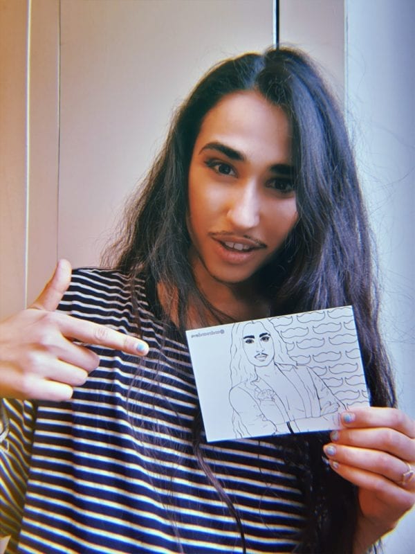 Christian Adore holds a postcard with the image of himself hand drawn in black and white on it. Christian is an olive skinned drag king with long black hair, black eyeliner and thin black moustache. He wears a black and white striped t shirt.