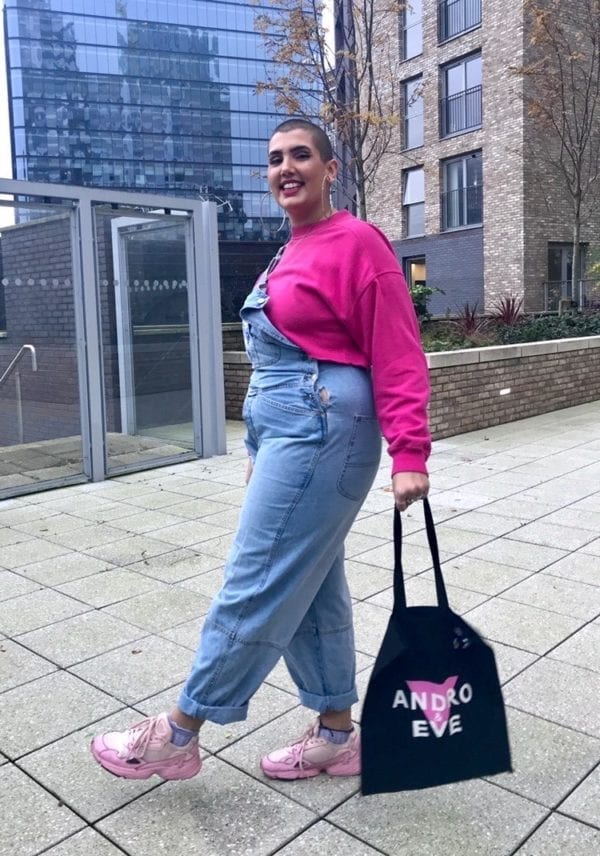 Genesia a Sotuh Asian woman with shaved head models the black logo tote with pink and white Andro and Eve logo. Sh wears denim dungarees and pink sweater.