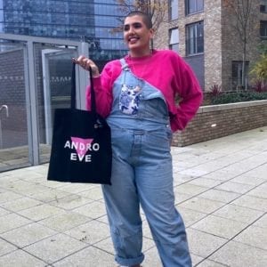 A woman with a shaved head smiles as she holds a black logo cotton tote bag, with pink and white design. She is wearing a bright pink long sleeved T shirt and blue denim dungarees and baby pink trainers. She stands outside in an urban area.