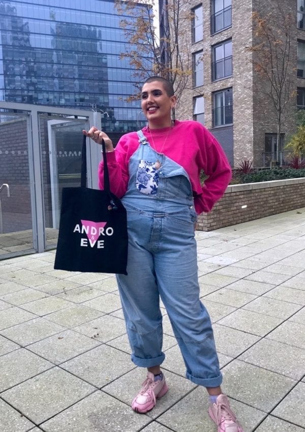 A woman with a shaved head smiles as she holds a black logo cotton tote bag, with pink and white design. She is wearing a bright pink long sleeved T shirt and blue denim dungarees and baby pink trainers. She stands outside in an urban area.