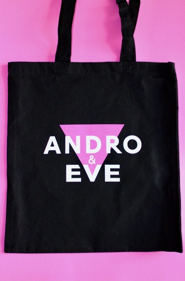 a black cotton tote bag with the trinagle of the andro and eve logo in pink and the words Andro and Eve overlaid in white.