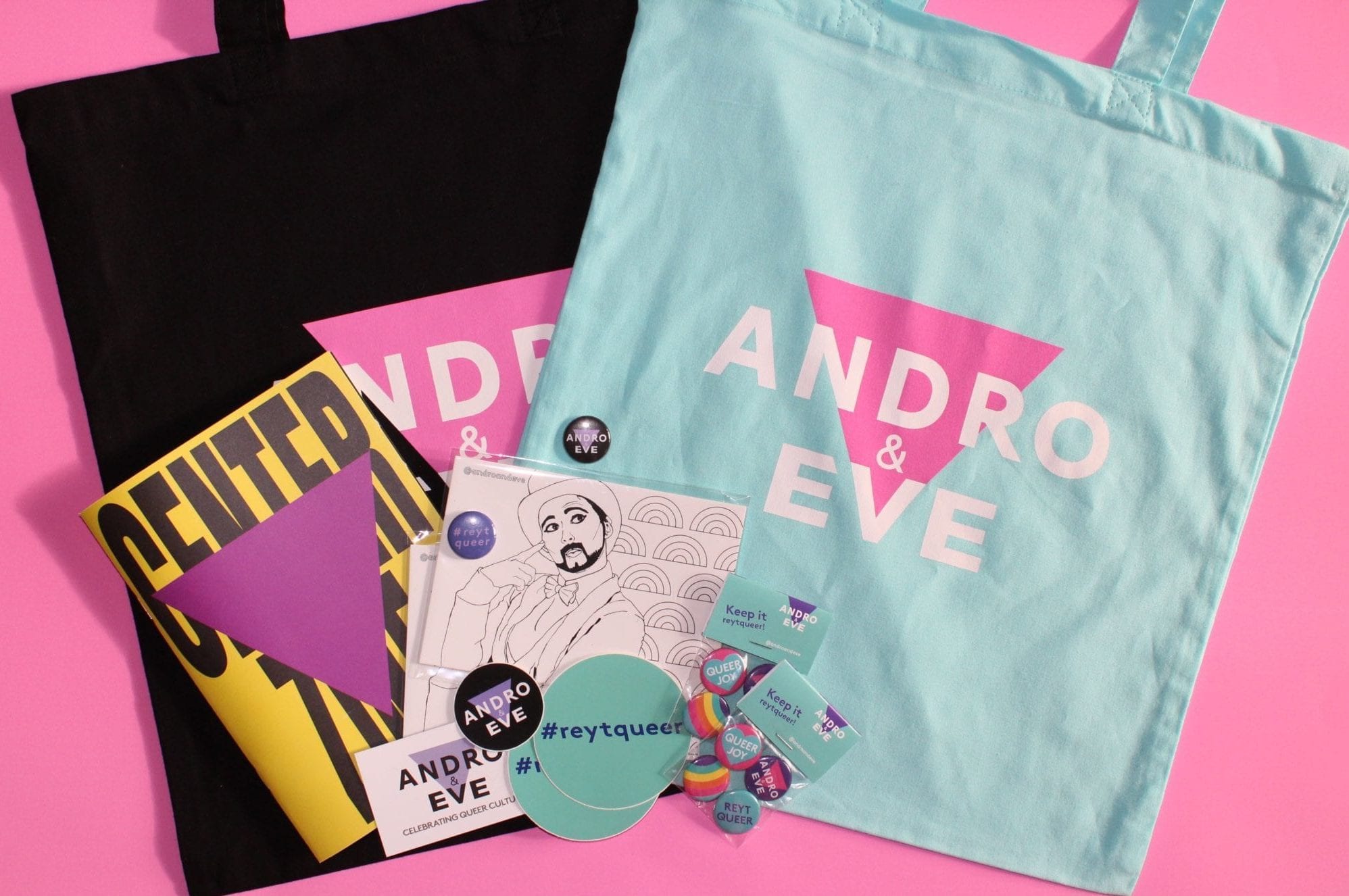 A turquoise and black cotton tote bag featuring the Andro and Eve logo sits underneath a pile of Andro and Eve merchandise. Including postcards, stickers and badges.