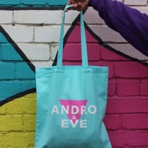 a turquoise tote bag is held by a hand against a painted wall in pink, turquoise and yellow. The tote bag has the Andro and Eve logo of inverted triangle overlaid with the words Andro and Eve, with ampersand printed in white.