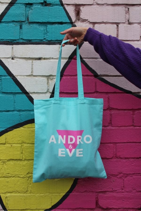 a turquoise tote bag is held by a hand against a painted wall in pink, turquoise and yellow. The tote bag has the Andro and Eve logo of inverted triangle overlaid with the words Andro and Eve, with ampersand printed in white.