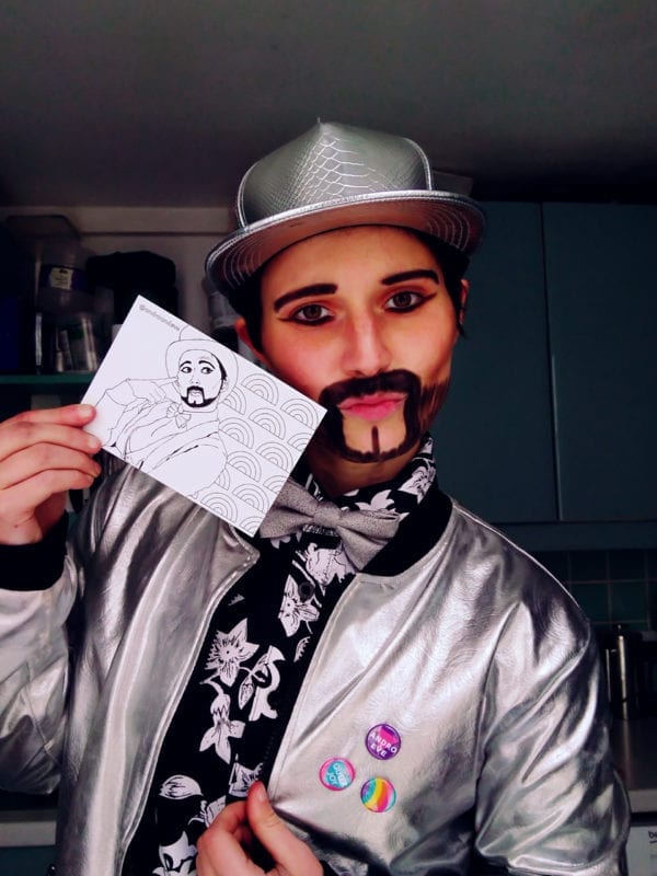 Drag King Sammy Silver holds a postcard featuring a hand drawn image of him in black and white. Sammy is a slim, white drag king with a heavy stylised beard and moustache. He wears a silver baseball cap, bow tie and bomber jacket and black patterned shirt.