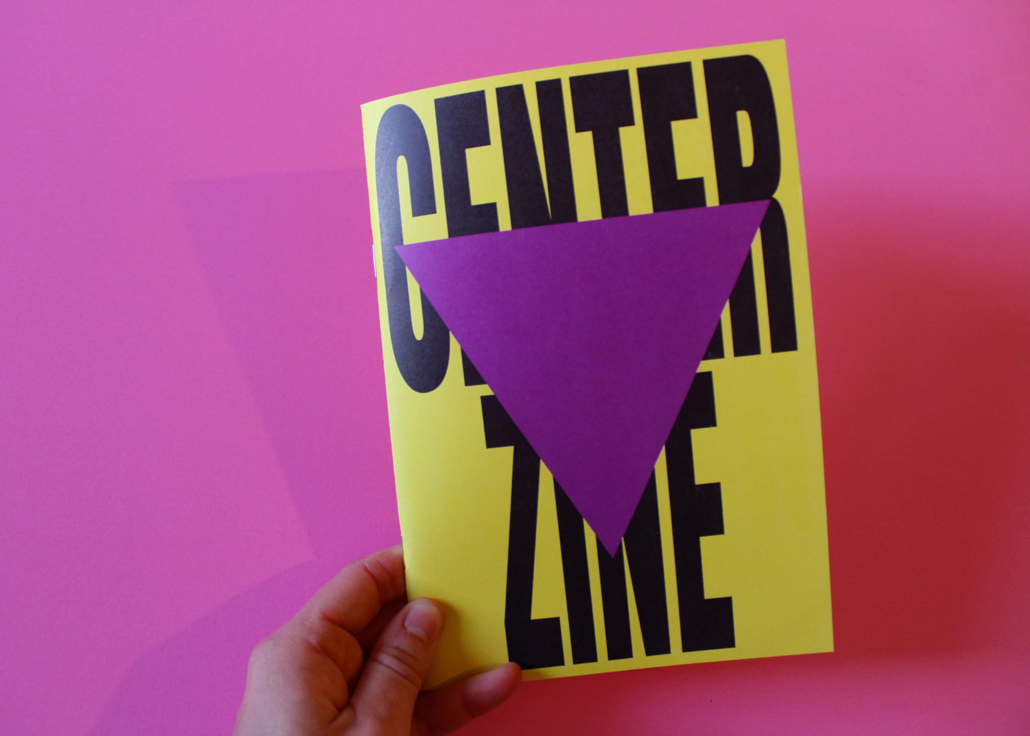 A white person's hand holds Centre zine. It has a bright yellow cover with bold black letters overlaid with a purple triangle. The text says 'Centre Zine'.