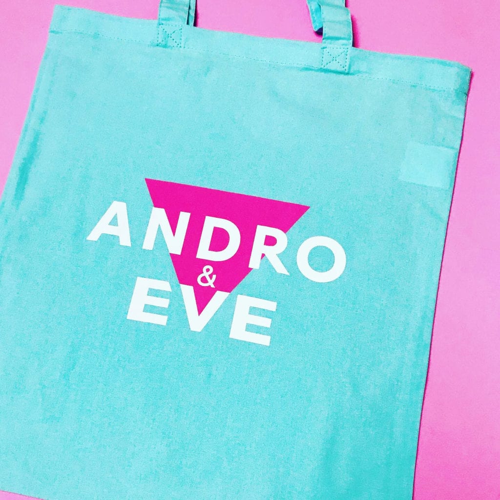 A pale turquoise cotton tote bag has the andro and eve logo printed on it in pink with white lettering. The design is in the centre of the bag. The bag sits on a pink surface.