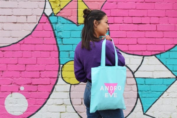 a slim, East Asian model with mid length black hair is wearing a purple jumper and holding a turquoise and pink andro and eve logo tote bag.