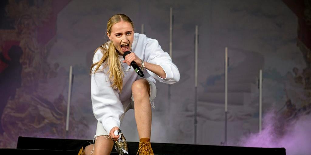 Silvana Imam, a slim, blonde haired rapper, kneels at the front of a stage rapping. She wears desert boots, shorts and white hoodie. 