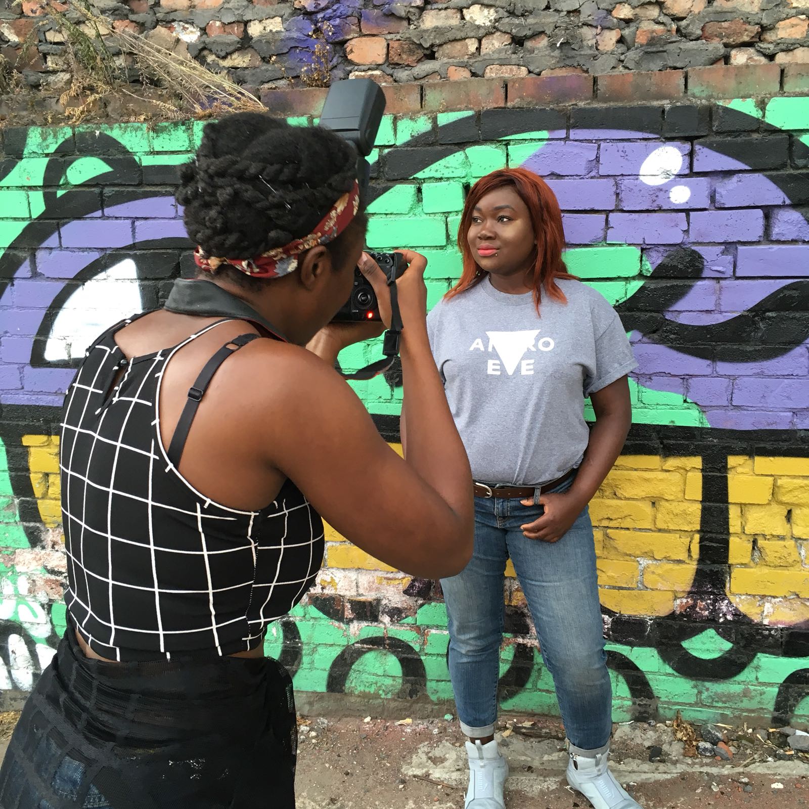 A Black, female photographer takes a picture of a Black, femme lesbian wwearing and Andro and Eve logo T Shirt