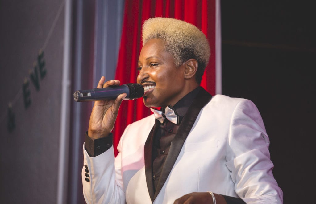 Drag king Don One stands on stage singing into a mic. He is a Black kind with short, bleach blonde hair, wwearing a white suit with black shirt and white bow tie. 