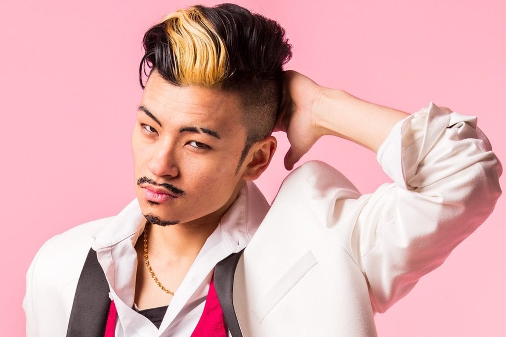 An east Asian drag king looks smoulderingly into the camera. He has short black quiff with a bleach blonde streak through it and shaved sides. He wears a white dinner jacket and ubuttoned white shirt with a shocking pink bow tie undone.