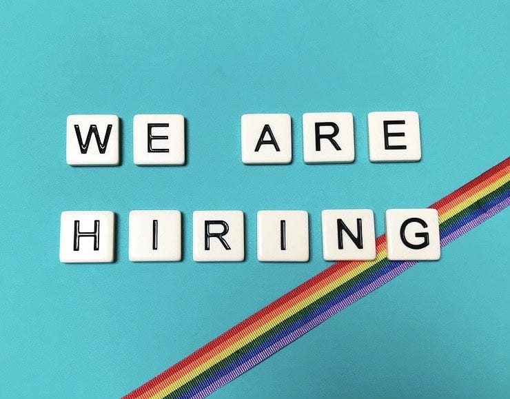 A turquoise tile has the words, we are hiring spelled out in scrabble tiles with a rainbow ribbon going across from top right corner to bottom left
