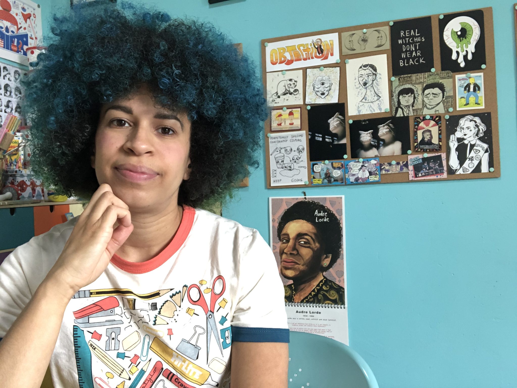 A Black woman with a turquoise afro sits at a desk with her hand under her chin. She is wearing a white T shirt with craft equipment illustrations on it. Behind her is a turquoise painted wall with a picture covered noticeboard on it