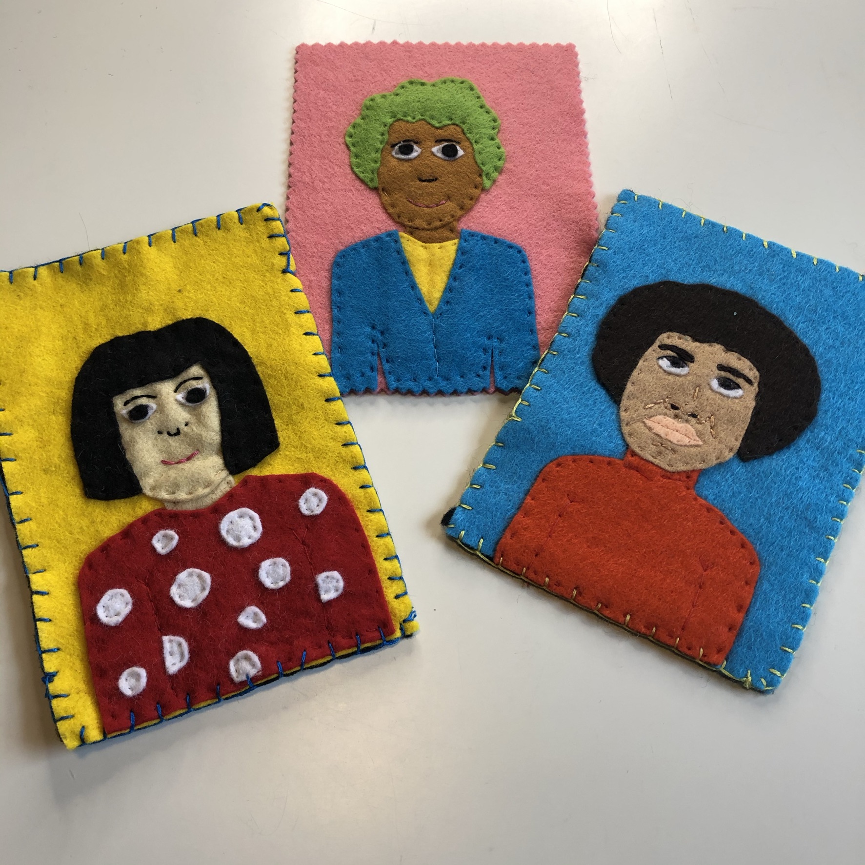 Three felt portraits in bright colours show a mix of people with different skin tones. The portraits are placed on a white table.