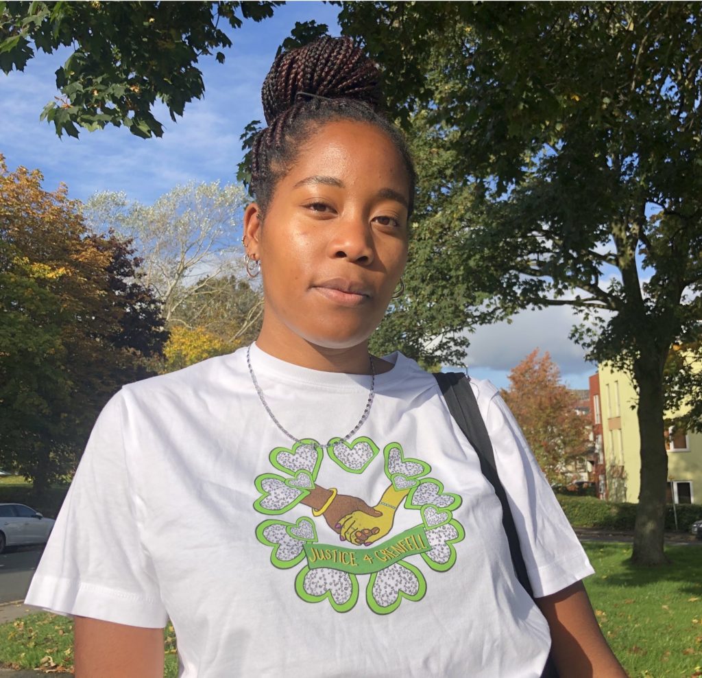 Image of a Black women who is staring into the camera with a slight smile, wearing a white t-shirt with print of green hearts surrounding, two hands held and writing that says, “JUSTICE 4 GRENFELL”. She is standing on the corner of the street, autumnal trees, cloudy blue skies, leaves on the grass and flats are in the background.