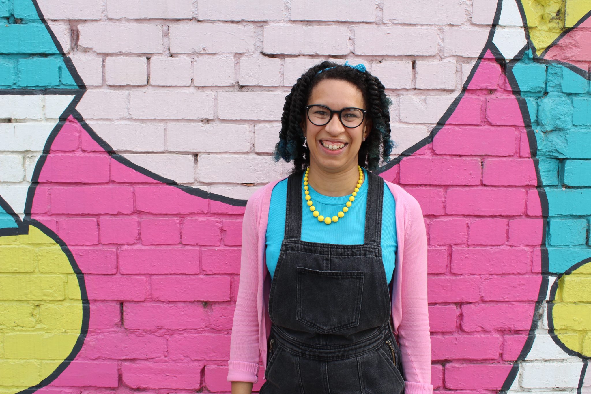 A Black woman with mid length hair stands in front a painted pink brick wall. She is wearing a turquoise tee with black dungarees and pink cardigan accessorised with bright yellow beaded necklace. She has on glasses and is smiling with an open smile at the camera