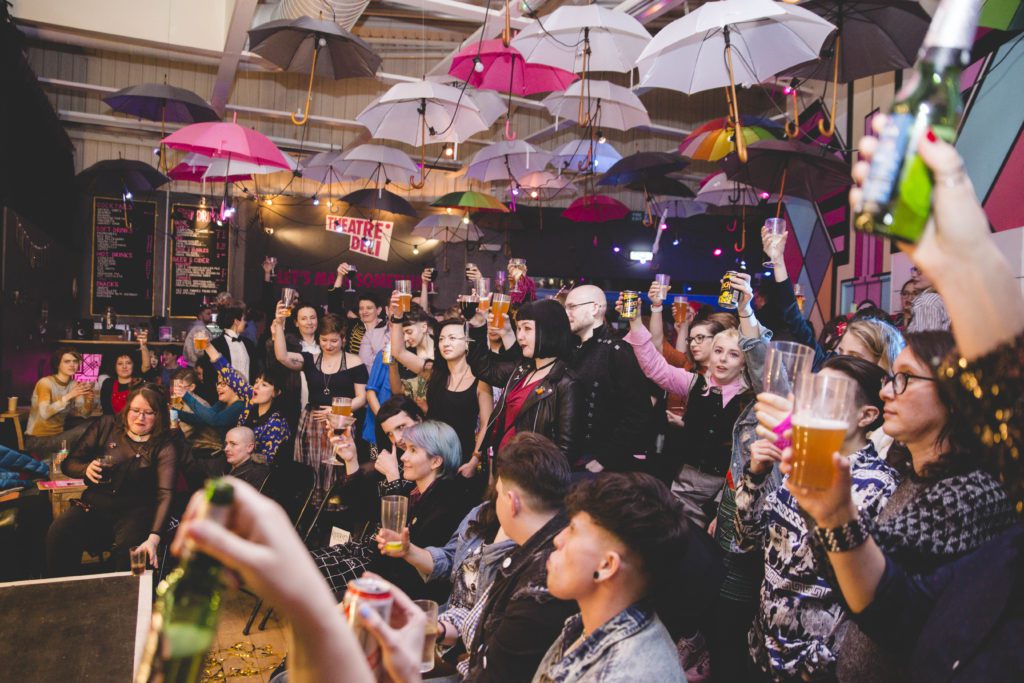 A crowd of people cheer on a performer out of shot. They are in a warmly lit venue with umbrellas above their heads. They have their hands raised clicking their fingers.