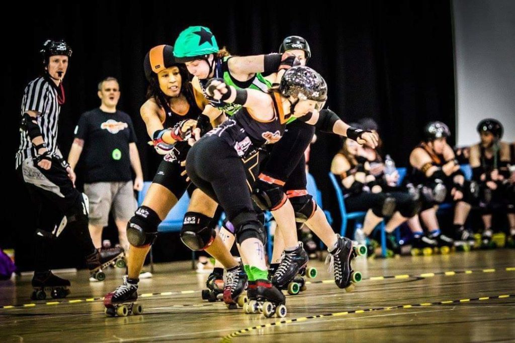 two roller derby players, wearing helmets and black close fitting outfits grapple with one another in an indoor gym.