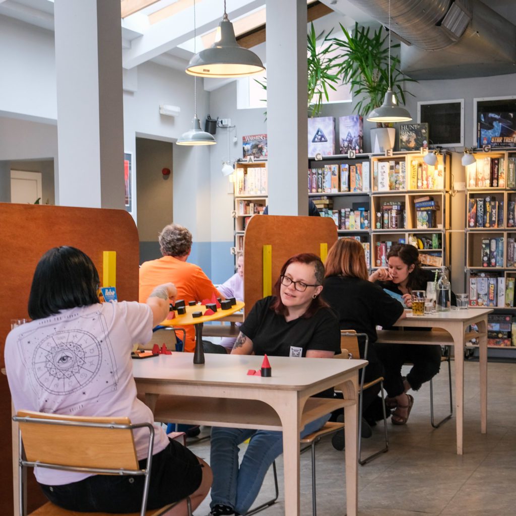 Two white people are shown in the foreground sat at a modern wooden table playing a board game. They are in a light and tal ceilinged cafe with pillars and a wall of board games in the background. 