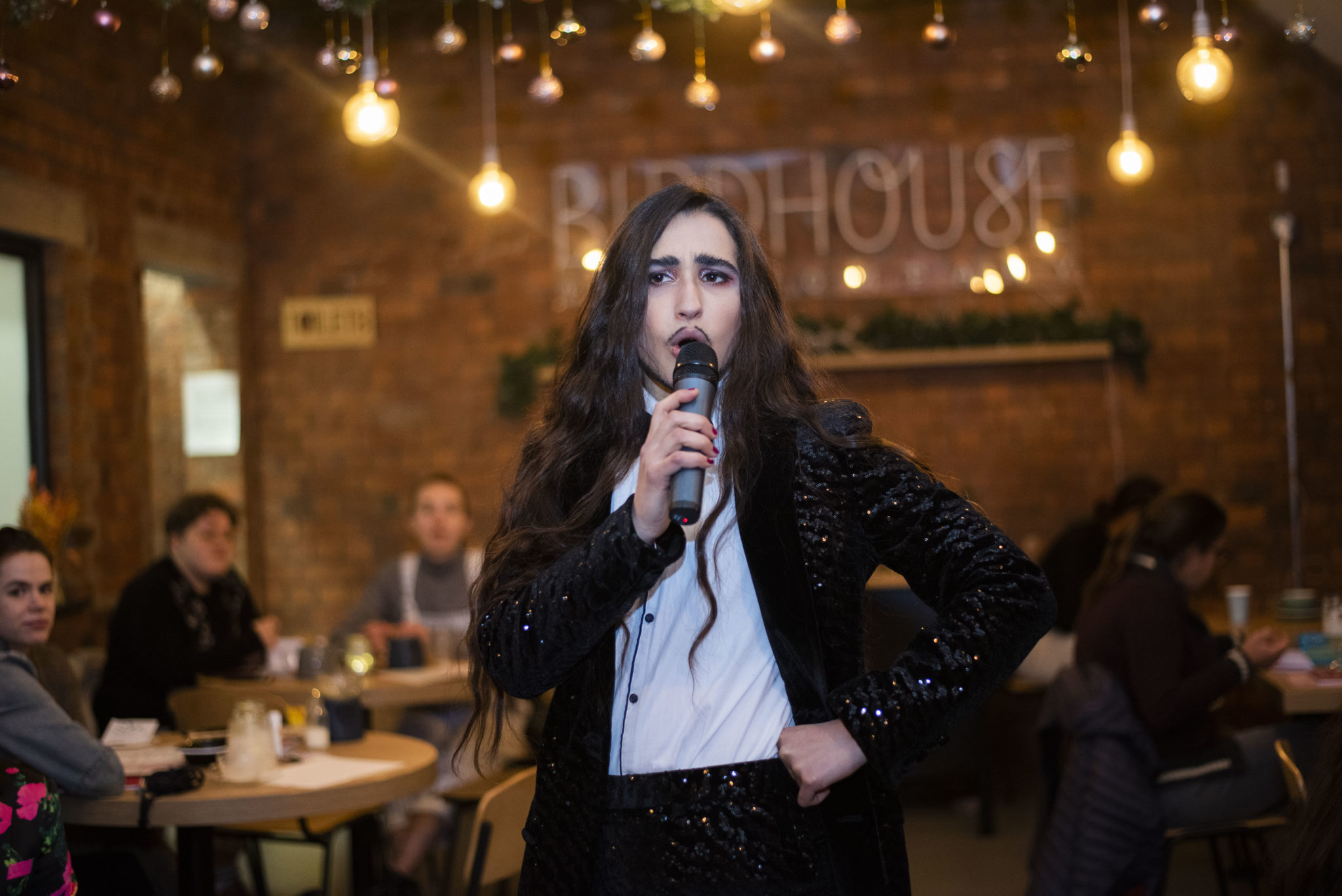 a slim, olive skinned drag king with brown, waist length hair and dark moustache sings into a microphone. He is wearing a sequin black suit with white shirt and bow tie, and smouldering eye shadow. He stands in a cafe with exposed brick walls with warm lighting.
