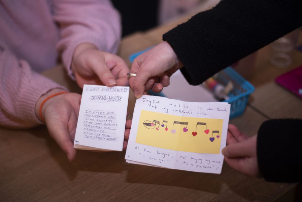 two sets of white hands hold up mini zines with drawings and text sharing joyful things