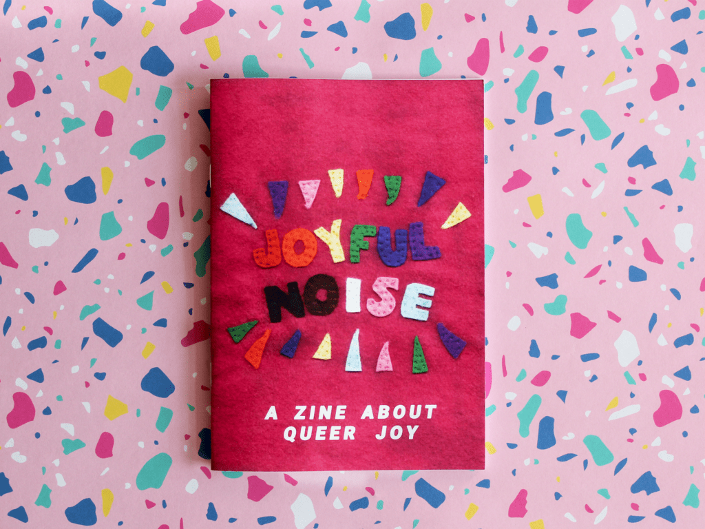 An A5 zine sits on a patterened pale pink background with a splodges of deep pink, blue, jade and yellow. The zine cover is a deep pink felt texture with the words ‘Joyful Noise’ stitched in multi coloured felt letters, below which white text states ‘ A zine about queer joy’.