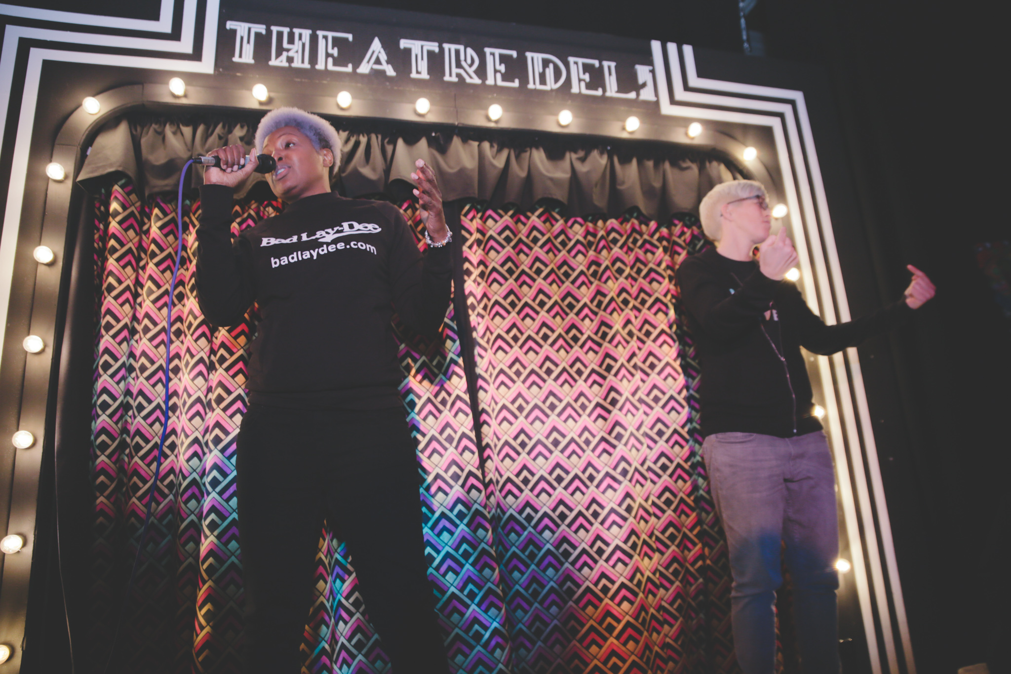 A Black, androgynous person stands on the right hand side of small stage speaking on a microphone. They are wearing a black tee with the words 'Bad Lay Dee' in white letters and black jeans. Their hair is short and bleached. On the right hand side of the stage is a BSL interpreter who is gesturing. She has cropped bleach blonde hair and is white. She wears a black hoodie and blue jeans. Behind the stage is a patterned pink curtain with warm white lightbulb surrounding, and Theatre Deli painted in white letters.