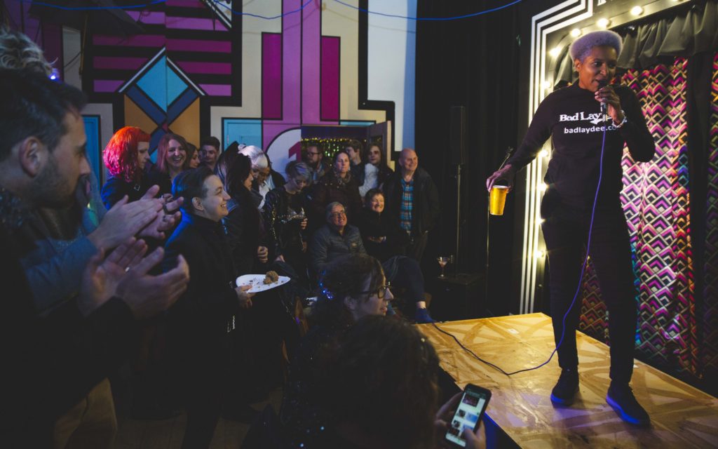 a Black androgynous rapper stands on a small stage holding a pint of beer while speaking into a mic. They have short bleached hair and wear black tee and jeans.A crowd of people seated and standing surrounds the small stage lit with warm lamps