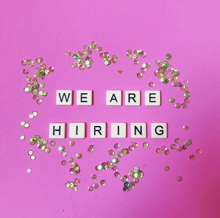 a set of cream tiles with black lettering spells out 'We are hiring'. The tiles are laid on a hot pink surface with iridescent sequins scattered around.