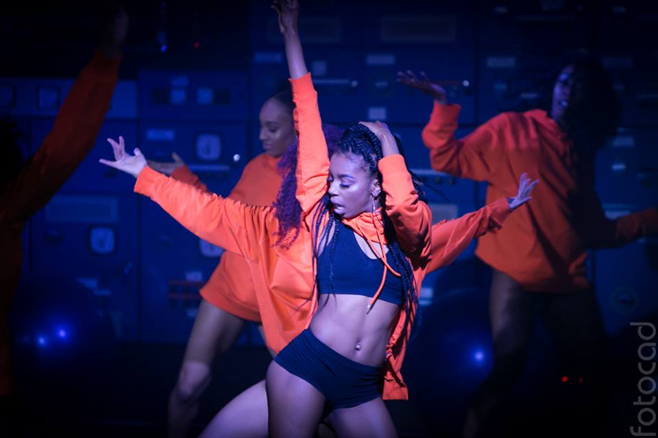 a group of Black, female and femme dancer strike poses onstage lit by blue lighting. They wear bright orange jackets and tight black dance wear.