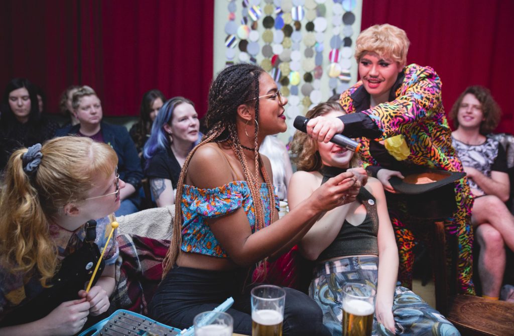 A Black femme presenting person with braids wearing an off the shoulder top is speaking into a microphone held by drag king Luke Warm. Luke, immersed in the audience of a cabaret is lit by pink lighting and he has short blonde hair and moustache and wears a colourful patterned suit