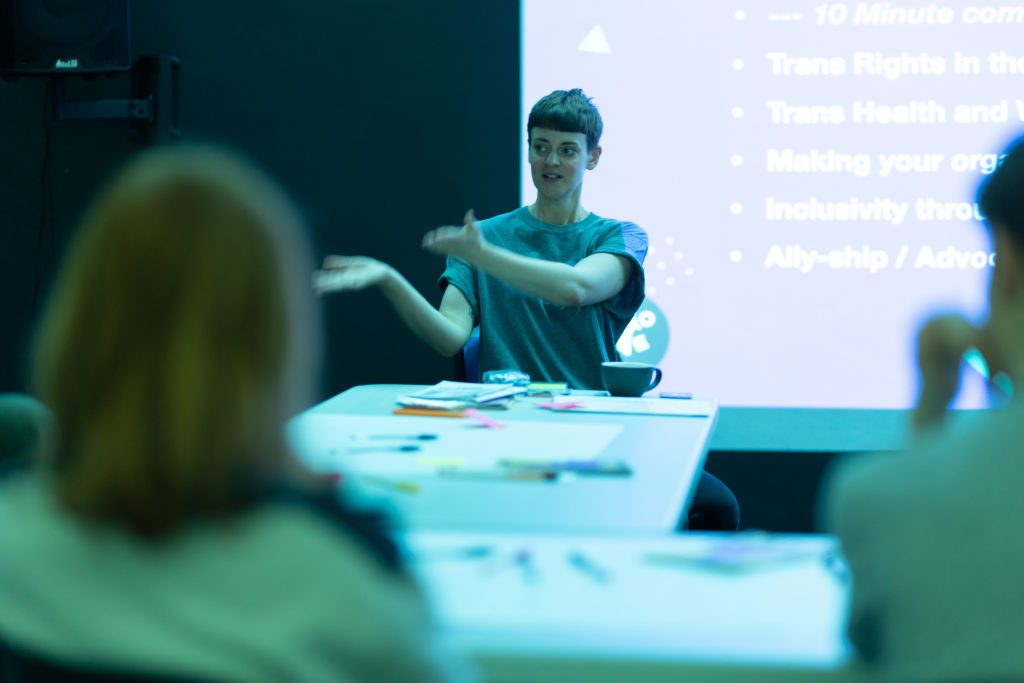 an androgynous white person with cropped brown hair sits at a table in front of a screen that has a projection it it. They have their arms outstretched in conversation and wear a grey Andro and Eve logo T shirt.