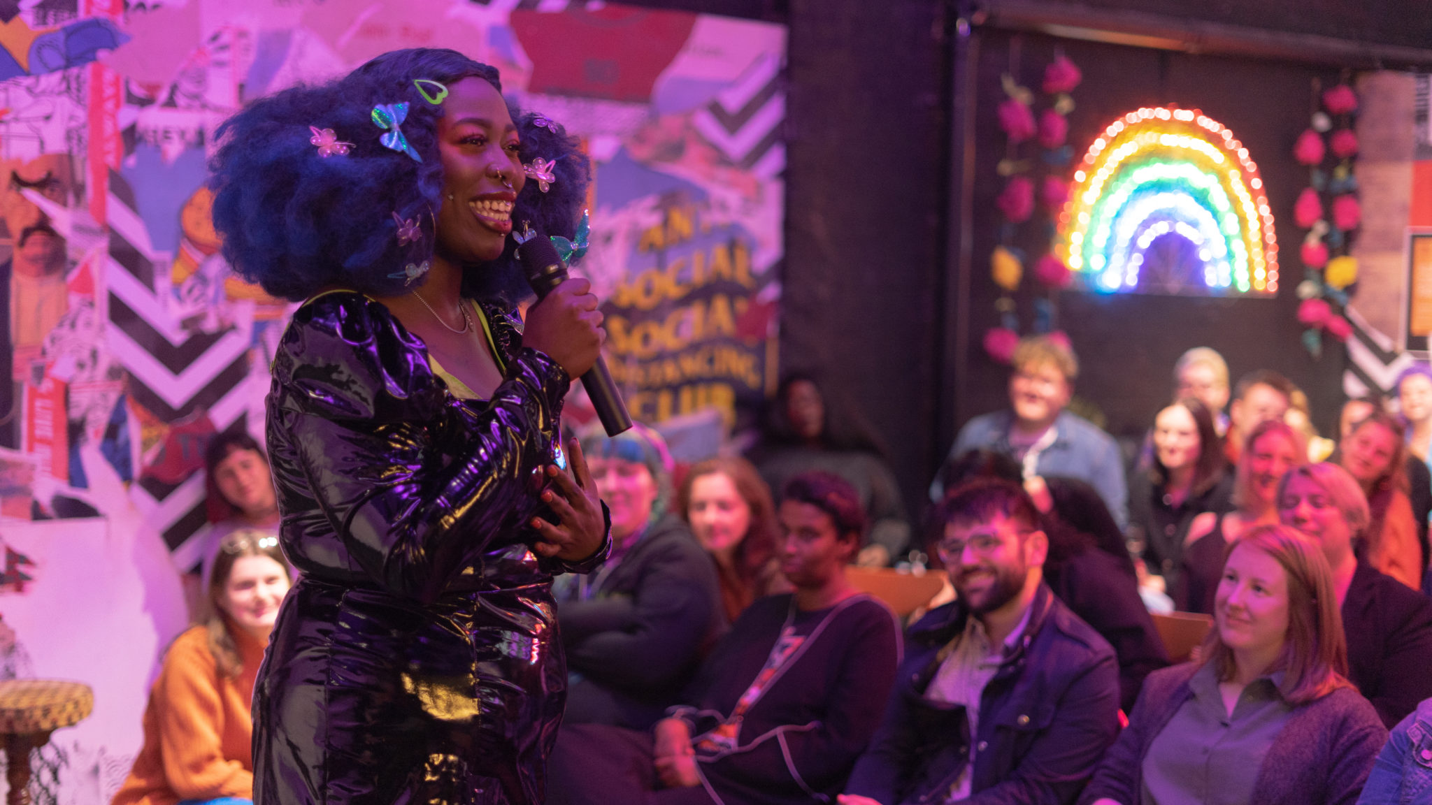 A black femme presenting person with blue Afro hair wears a black PVC mini dress and smiles on a mix in front of a crowd of people. She is lit by purple lighting.
