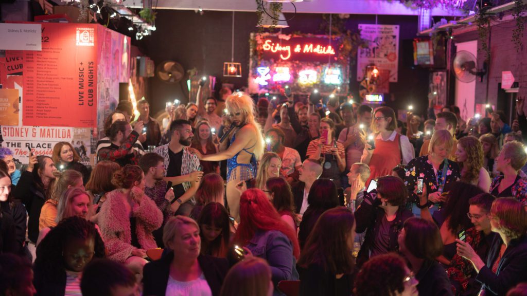 A drag performer with a blonde wig stands in the middle of a crowd of people in a bar with everyone in the crowd having their phone camera lights shining 