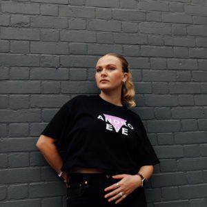 A white woman with blonde hair models a black logo tee in natural light. She is wearing black jeans and the logo is a lilac triangle overlaid with white letters that say Andro & Eve