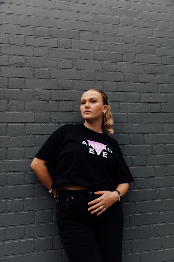 A white woman with blonde hair models a black logo tee in natural light. She is wearing black jeans and the logo is a lilac triangle overlaid with white letters that say Andro & Eve